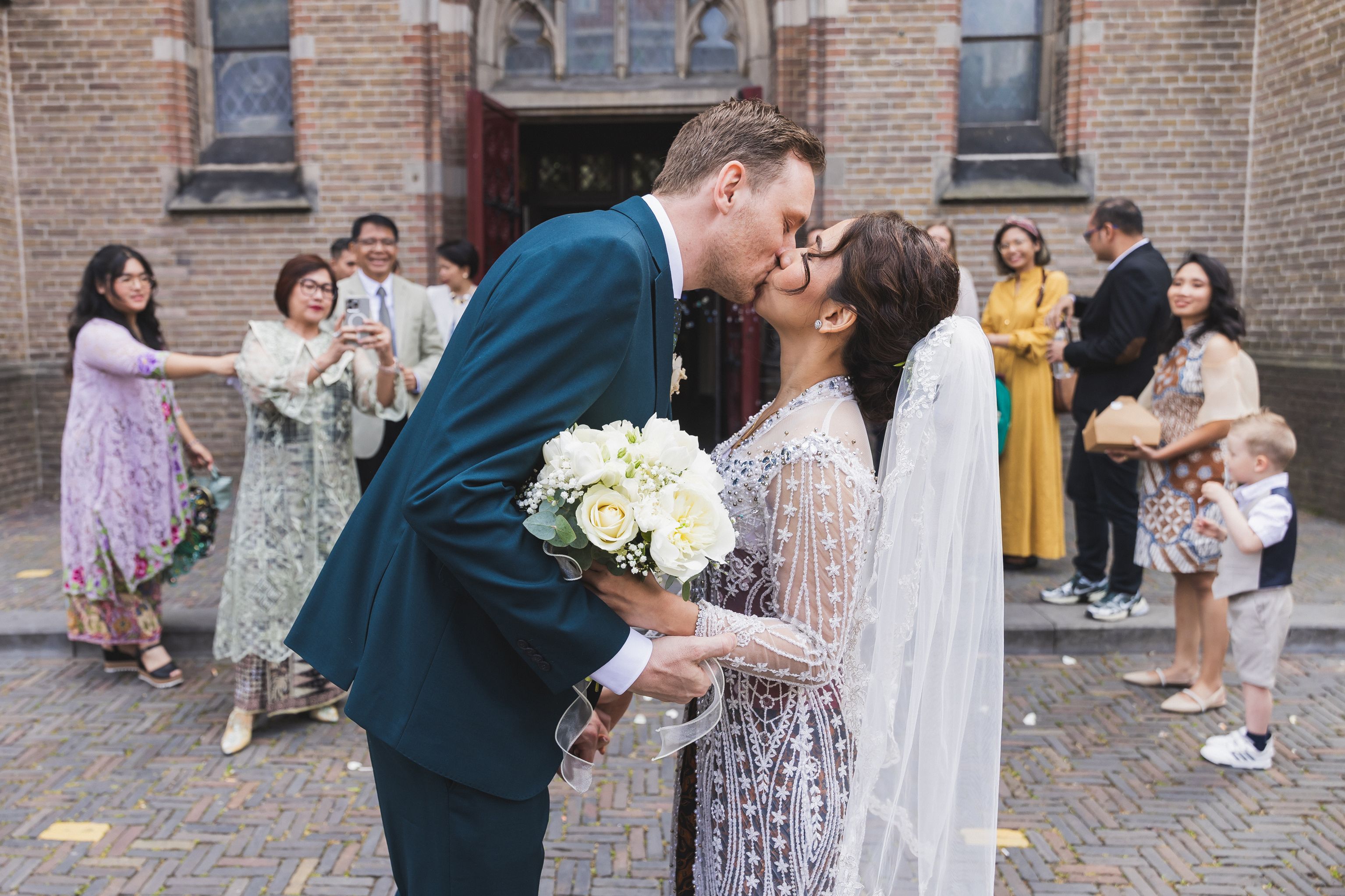 Romantische trouwfotografie van bruidspaar dat kust voor een kerk, met gasten op de achtergrond. Joep van Drunen Photography