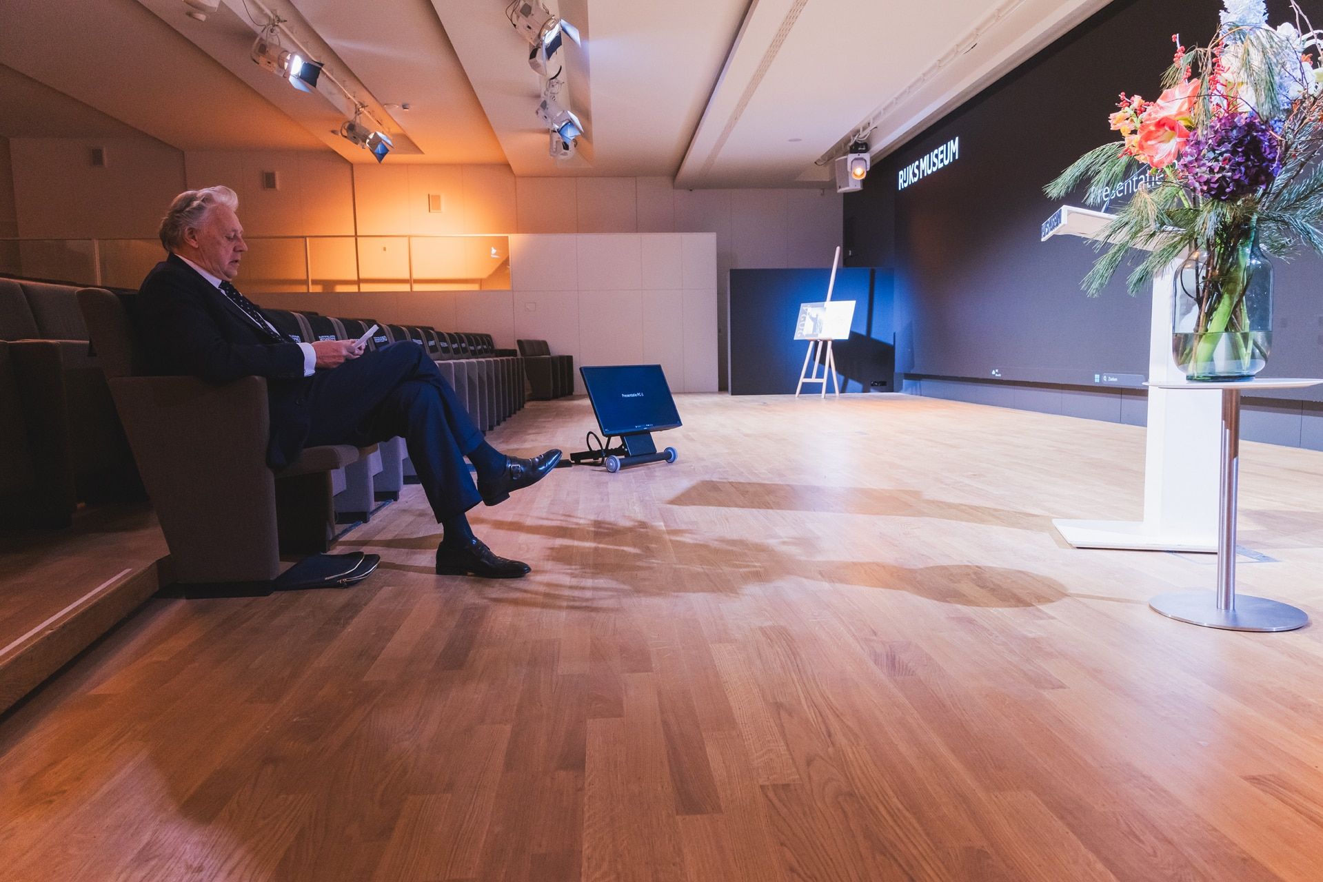 Zaal voor Abe Bonnema Prijs uitreiking, man leest document. Podium met bloemen en scherm met 'Rijksmuseum'. JVD_8746.jpg Joep van Drunen Photography