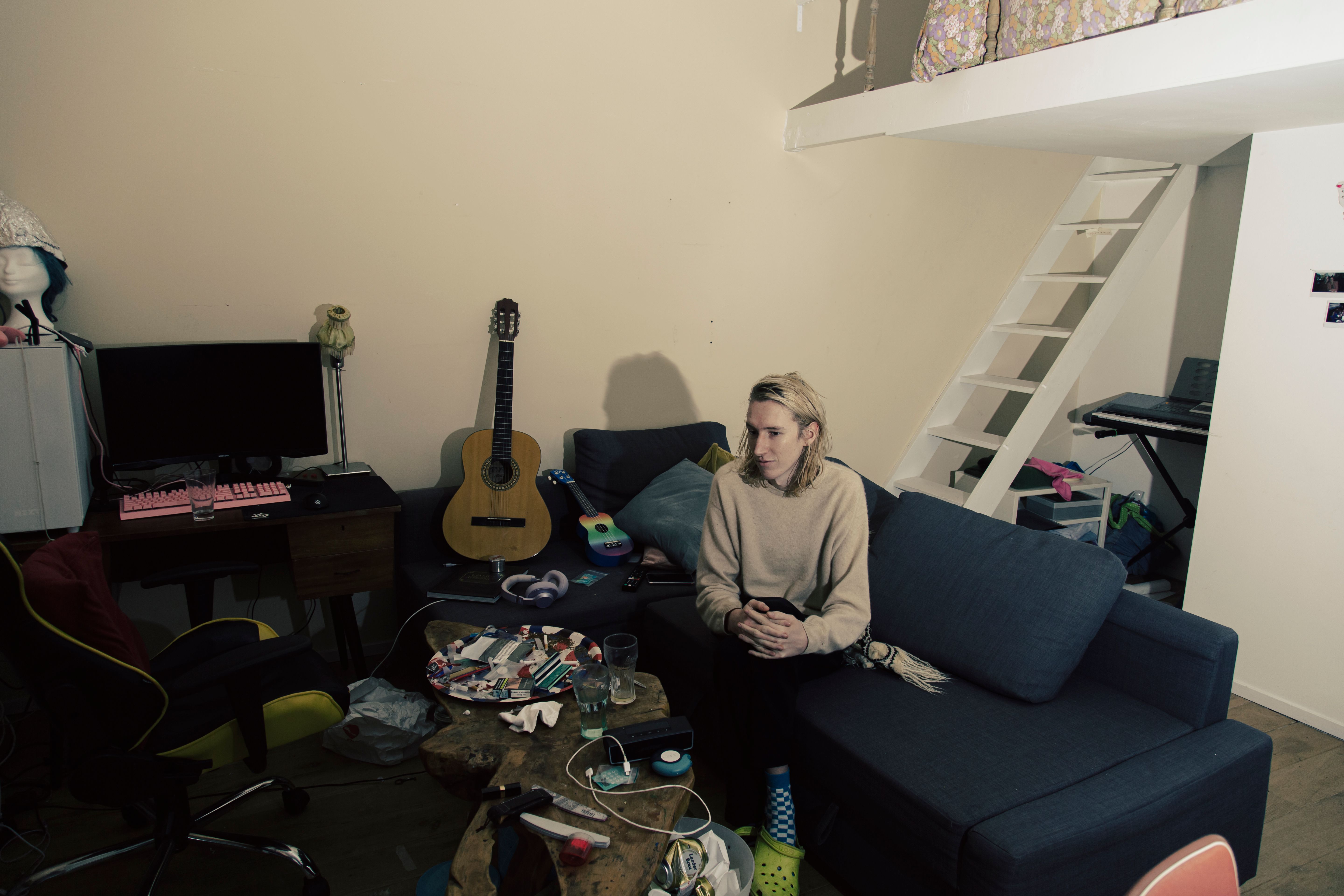 Senna Bodile zit op een bank in een kamer met gitaar, computer en trap naar een bedstee. Joep van Drunen Photography