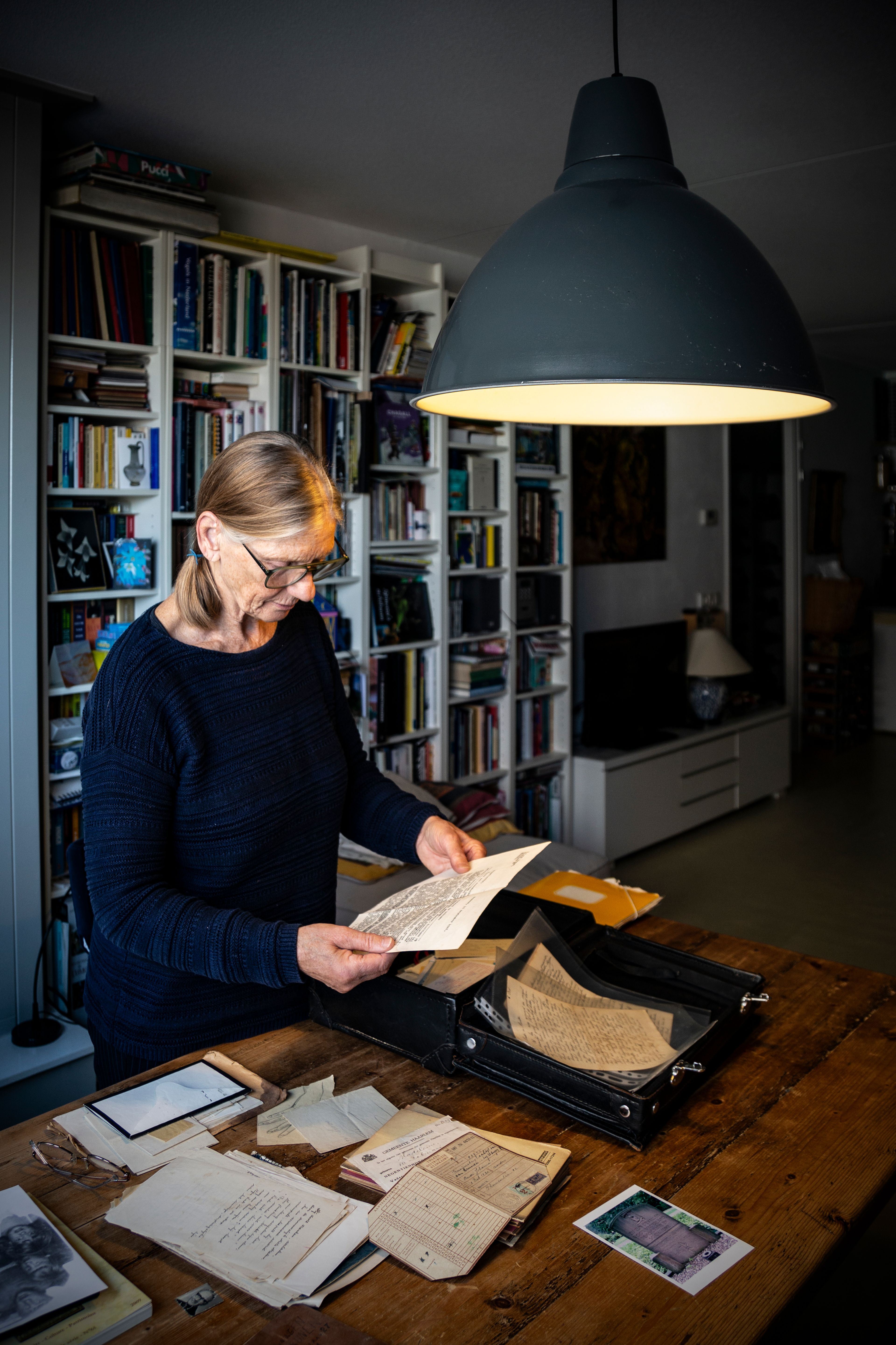 Madeleine Boek leest documenten aan tafel. Karel Jan onderzoek. Interieur met boekenkast en lamp. Joep van Drunen Photography