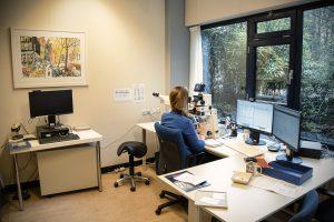 Documentaire autonoom: Vrouw werkt met microscoop in een lichte kamer met computers en een schilderij aan de muur. Joep van Drunen Photography