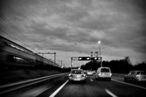 Zwart-wit foto van een trein naast de snelweg, documentaire stijl. Autonoom verkeer met auto's onder een bewolkte lucht. Joep van Drunen Photography