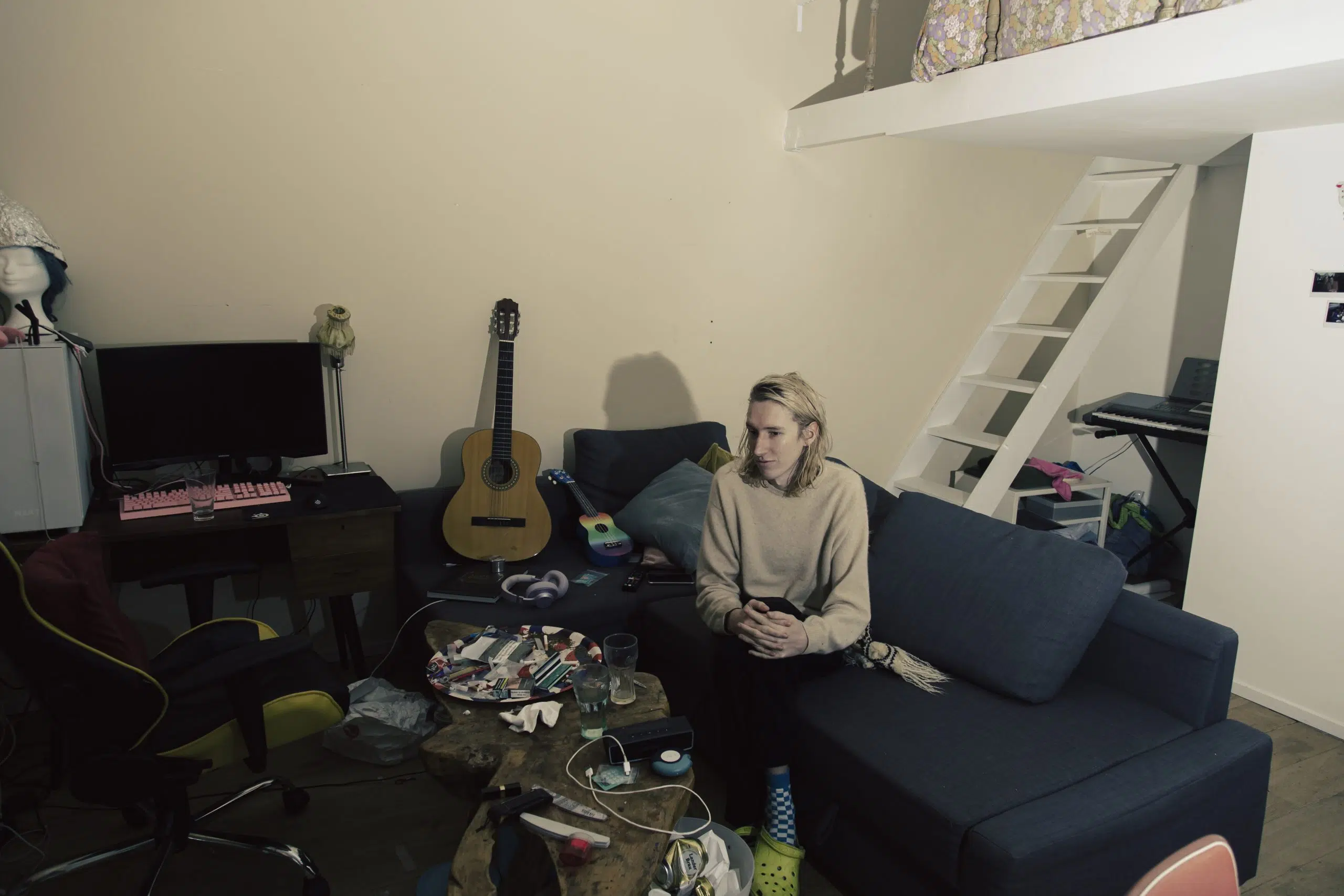 Autonoom persoon zit op een bank in een rommelige kamer met gitaar en bureau. Documentaire foto. Joep van Drunen Photography