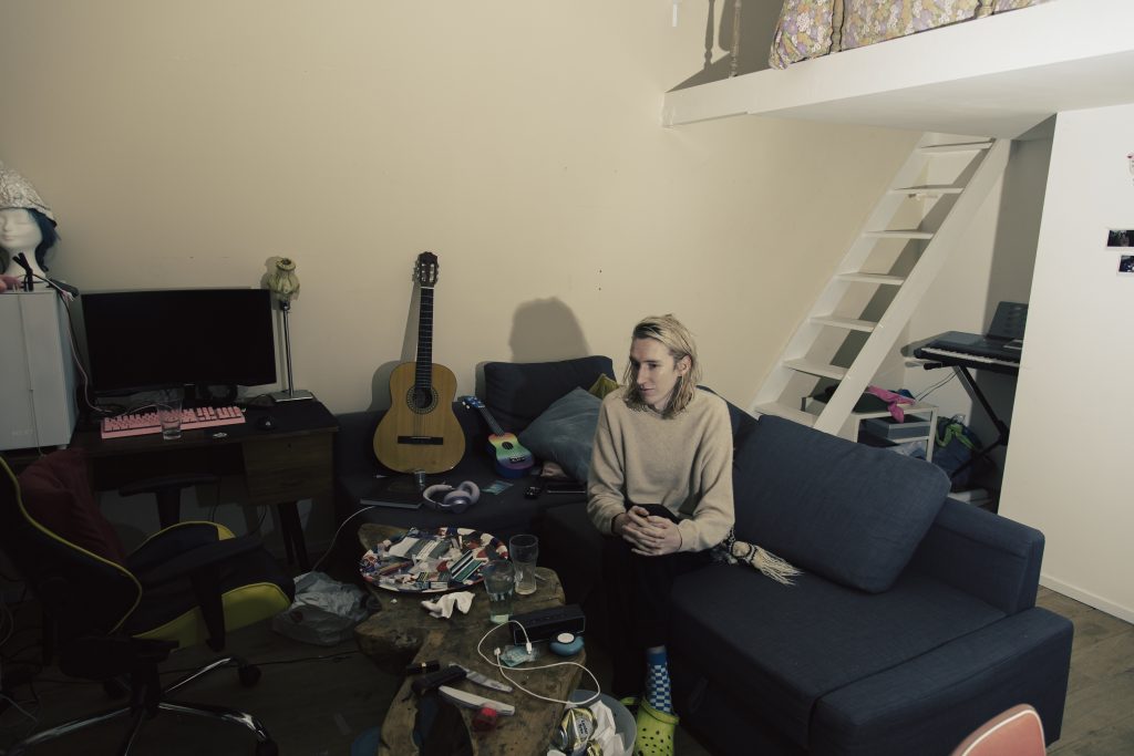 Senna Bodile zit op een bank in een kamer met gitaar, computer en trap naar een bedstee. Joep van Drunen Photography