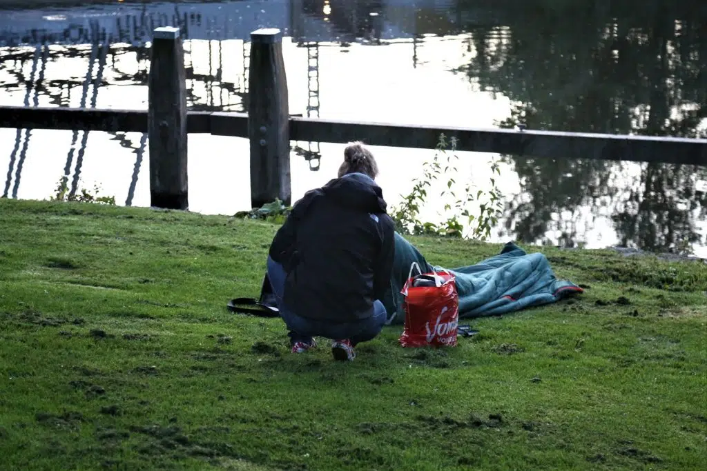 Veldwerk foto: Persoon bij een slaapzak aan de waterkant. Groen gras en een rode tas. Joep van Drunen Photography