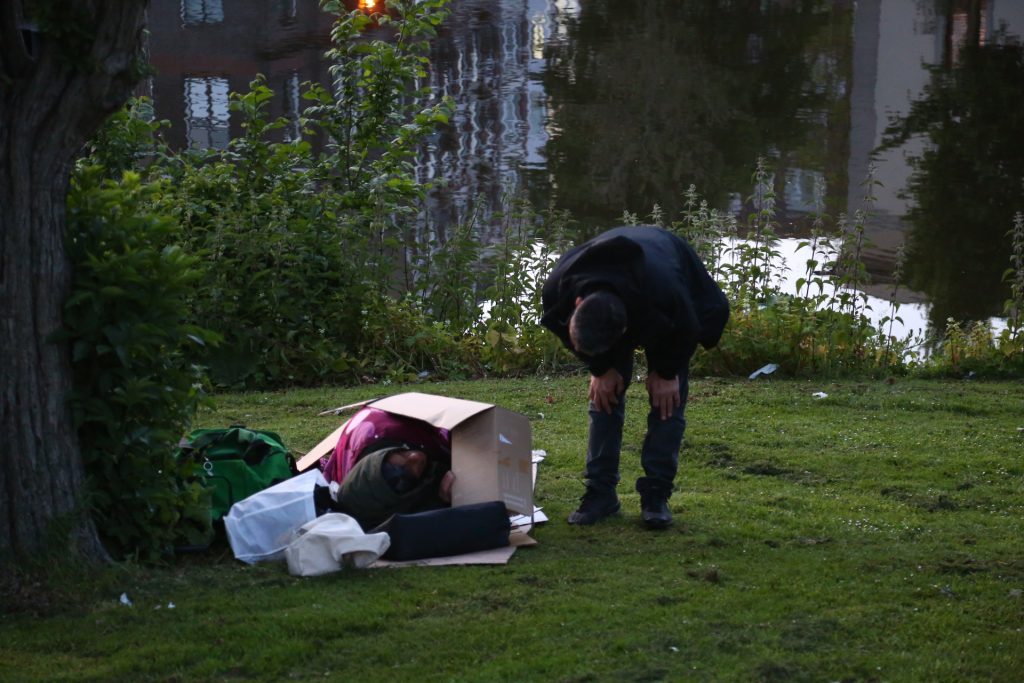 Veldwerk: Man buigt zich voor kartonnen doos met slapende persoon op grasveld. Joep van Drunen Photography