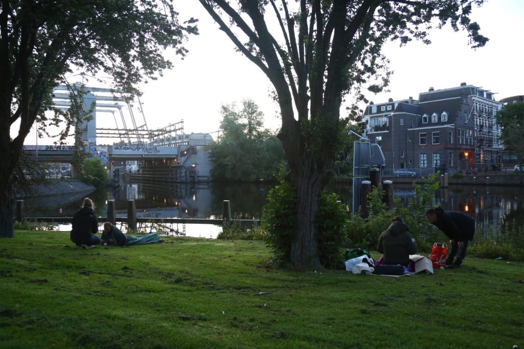 Veldwerk aan het water met brug en gebouwen op de achtergrond. Groep mensen ontspant in het gras. Joep van Drunen Photography