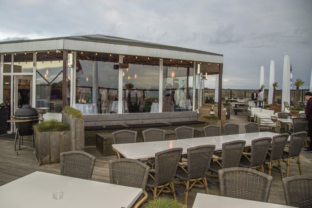Buitenaanzicht van een strandtent met tafels en stoelen op een houten terras, klaar voor bezoekers. Joep van Drunen Photography