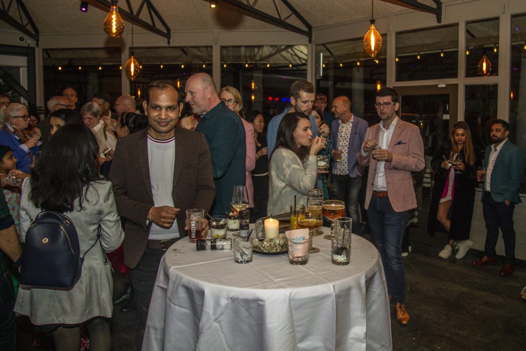 Groep mensen op evenement, IS5A3999, met drankjes rond een tafel. Sfeervolle verlichting. Joep van Drunen Photography