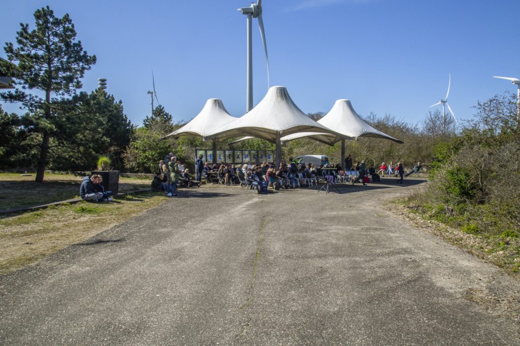 Bijeenkomst in het Deltapark met windmolens op de achtergrond. Mensen zitten onder een overkapping. IS5A4243. Joep van Drunen Photography