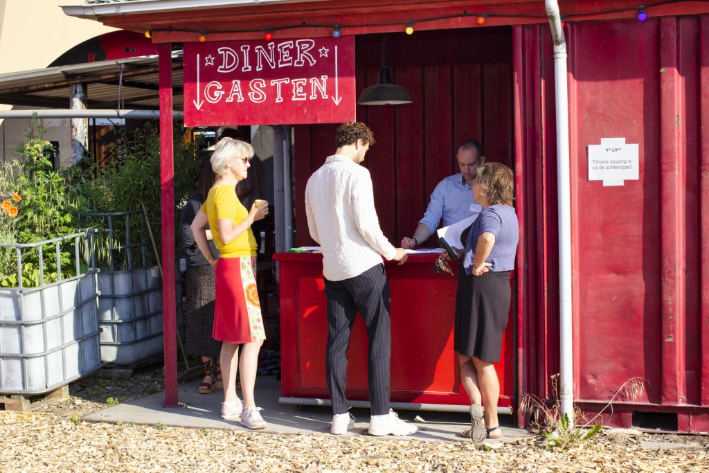 Mensen bij een rode 'Diner Gasten' kraam, mogelijk een evenement. IMG_1895.jpg. Joep van Drunen Photography