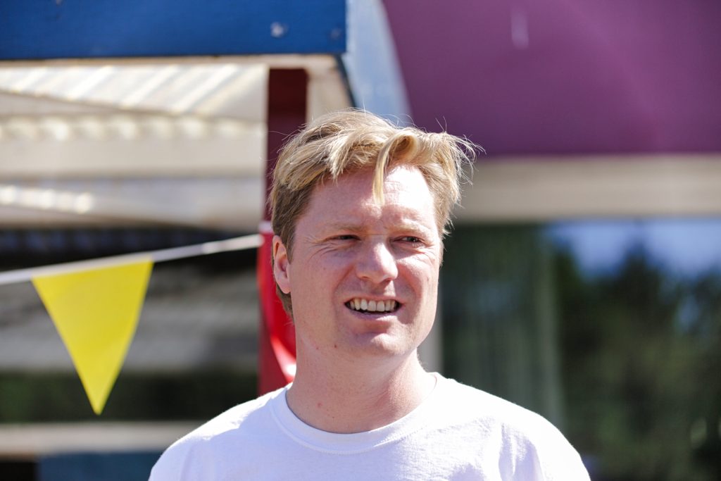 Portret van man met blond haar buitenshuis, lachend en gekleed in een wit T-shirt. Achtergrond met gele vlaggetjes. Joep van Drunen Photography