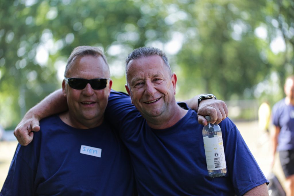 Twee lachende mannen, vriendschappelijk omhelsd, één met zonnebril en naamsticker, de ander met een flesje water. Joep van Drunen Photography